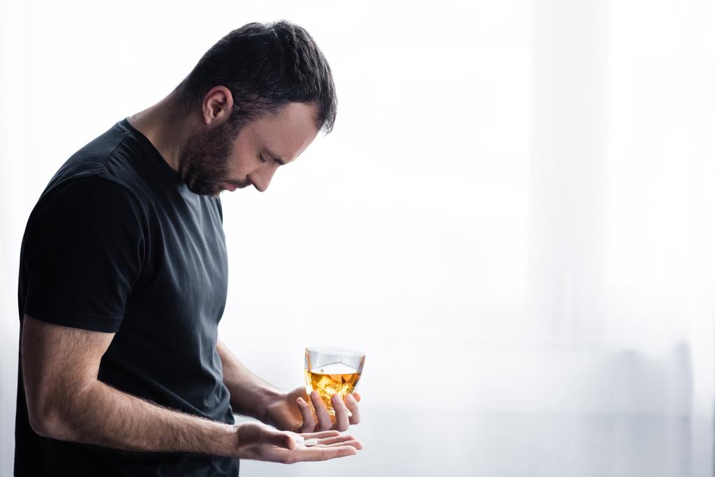 depressed man in black t-shirt standing with glass of whiskey and handful of pills - Photo, Image