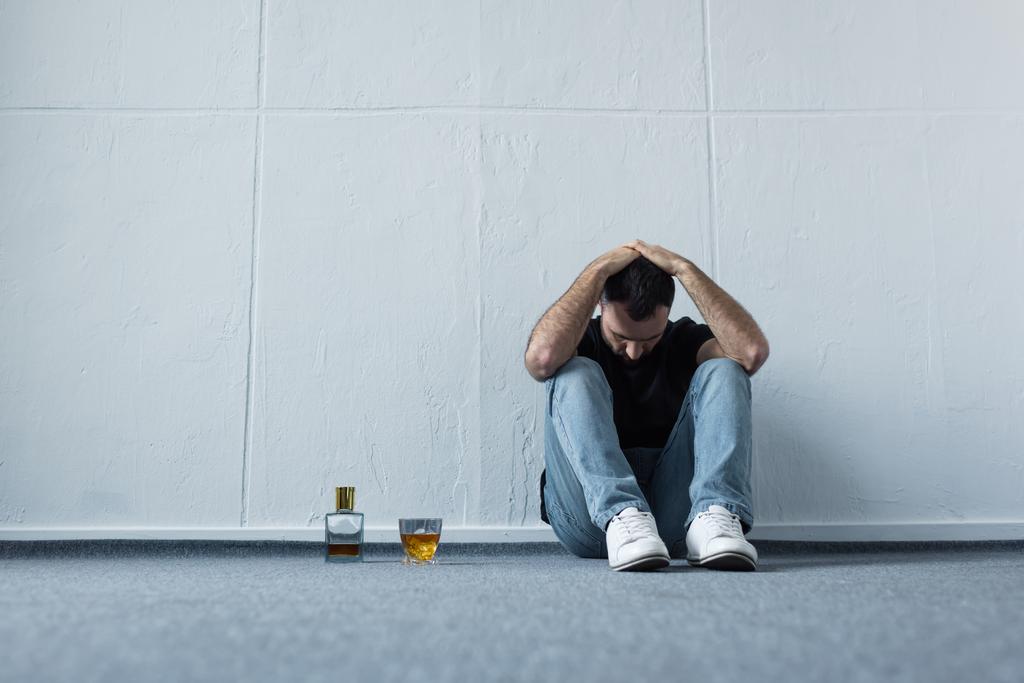 подавленный мужчина сидит на полу у белой стены возле бутылки и стакана виски
 - Фото, изображение