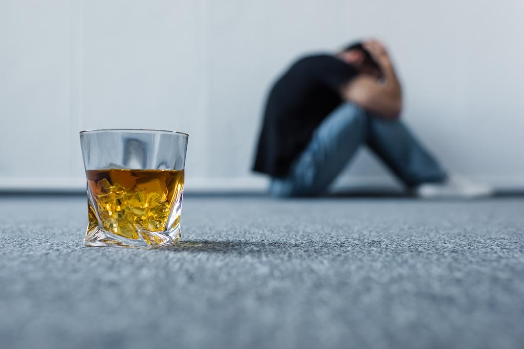 επιλεκτική εστίαση του καταθλιπτικού ανθρώπου που υποφέρει ενώ κάθεται σε γκρίζο πάτωμα κοντά σε ποτήρι ουίσκι - Φωτογραφία, εικόνα