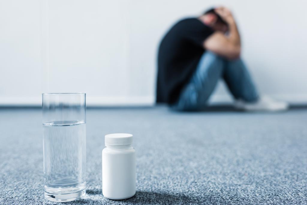 επιλεκτική εστίαση του καταθλιπτικού ανθρώπου που υποφέρει ενώ κάθεται στο πάτωμα από τοίχο κοντά σε κοντέινερ με χάπια και ποτήρι νερό - Φωτογραφία, εικόνα