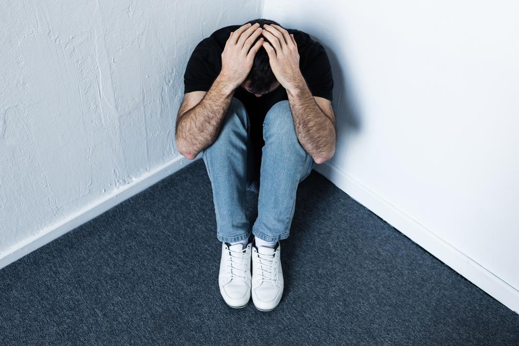 депресивний чоловік сидить на сірій підлозі в кутку і тримає руки на голові
 - Фото, зображення