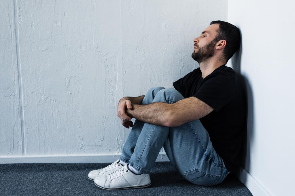 knappe depressieve man zittend op de vloer in de hoek door witte muur en op zoek weg - Foto, afbeelding