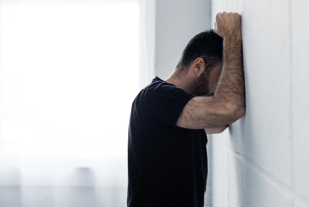 erwachsener depressiver Mann in schwarzem T-Shirt leidet, während er an weißer Wand steht - Foto, Bild
