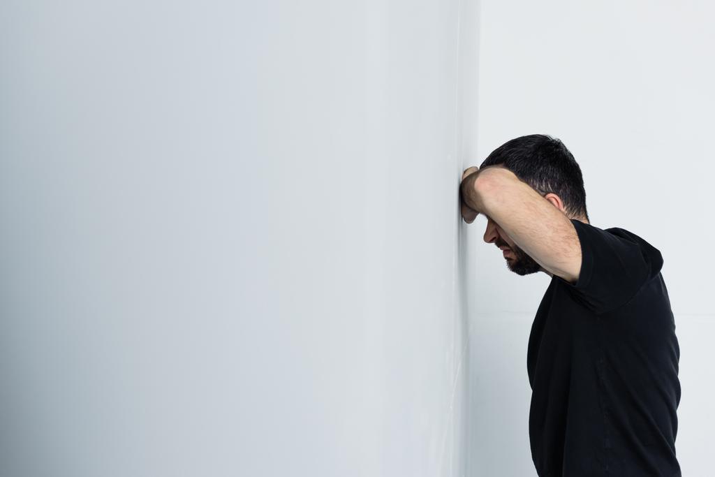 ενήλικος αναστατωμένος άνθρωπος με μαύρο μπλουζάκι που στέκεται από λευκό τοίχο  - Φωτογραφία, εικόνα