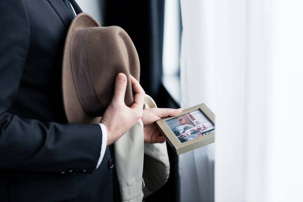 обрізаний вид людини, що тримає фотографію в кадрі, стоячи біля вікна з капелюхом в руці
 - Фото, зображення