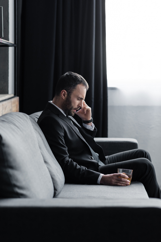 Aufgebrachter Geschäftsmann im Anzug sitzt mit Glas Whiskey auf Sofa und benutzt Smartphone - Foto, Bild