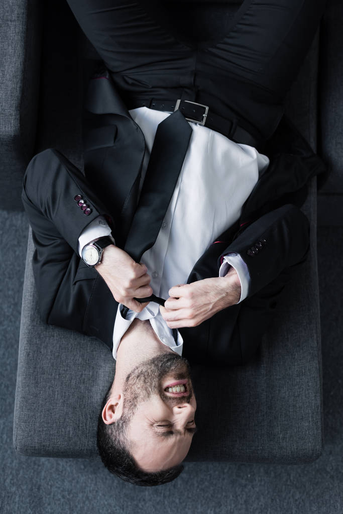 Draufsicht des depressiven Geschäftsmannes im schwarzen Anzug, der auf dem Sofa leidet  - Foto, Bild