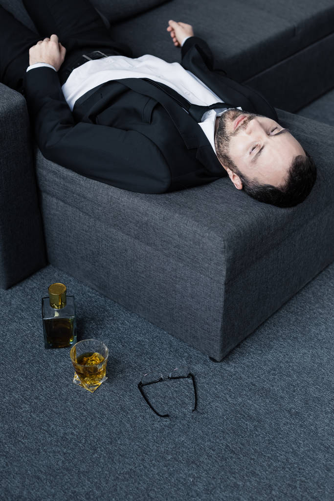 床の上のメガネ、ボトル、ウイスキーのグラスの近くにソファに横たわっている動揺したビジネスマンの高い角度の眺め - 写真・画像