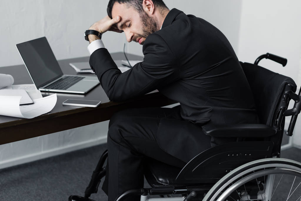 homme d'affaires handicapé déprimé en fauteuil roulant assis sur le lieu de travail avec ordinateur portable, smartphone et papiers
 - Photo, image