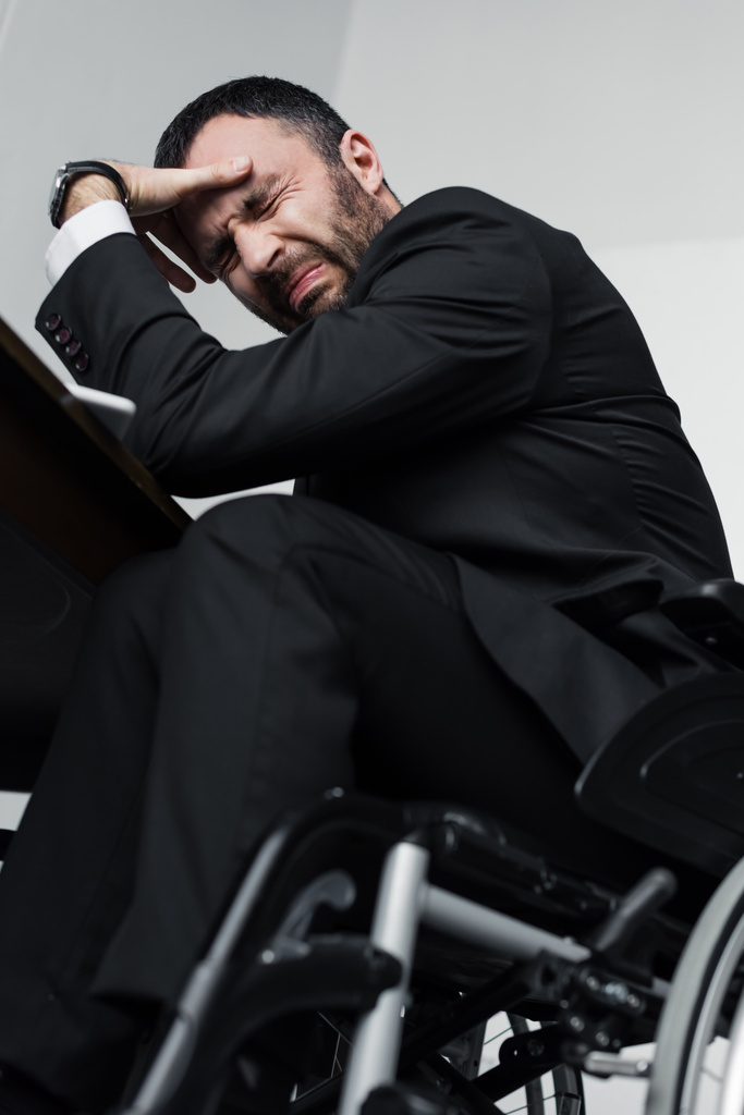 χαμηλή γωνία θέα της καταθλιπτικός επιχειρηματίας κλαίει ενώ κάθεται σε αναπηρικό αμαξίδιο - Φωτογραφία, εικόνα