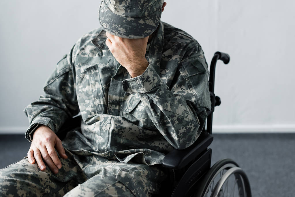 deprimovaný tělesně postičený muž v vojenské uniformě sedící na invalidním vozíku a držící ruku na obličeji - Fotografie, Obrázek