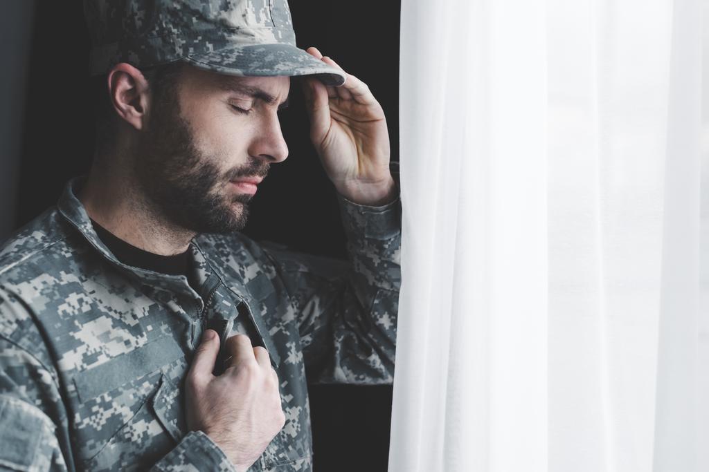nachdenklicher bärtiger Mann in Uniform, der die Hand auf der Brust hält, während er mit geschlossenen Augen am Fenster steht - Foto, Bild