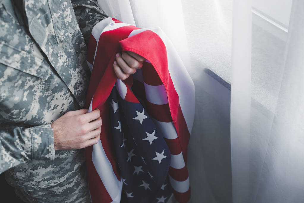 Ausgeschnittene Ansicht eines Mannes in Militäruniform, der am Fenster eine US-Nationalflagge hält - Foto, Bild