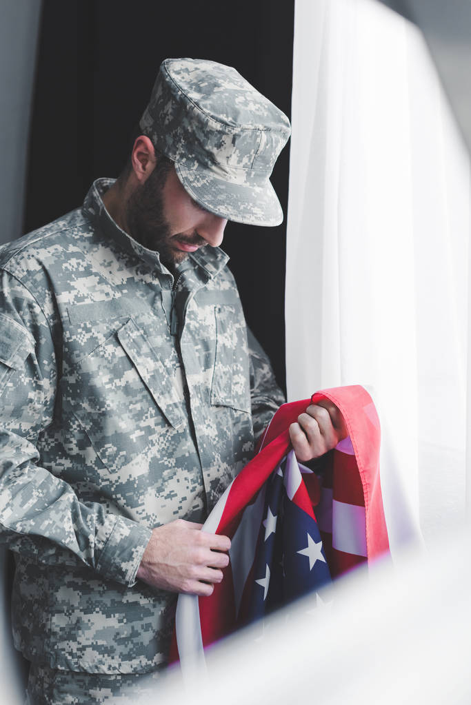 messa a fuoco selettiva di triste militare in uniforme che tiene USA bandiera nazionale mentre in piedi vicino alla finestra con la testa piegata
 - Foto, immagini
