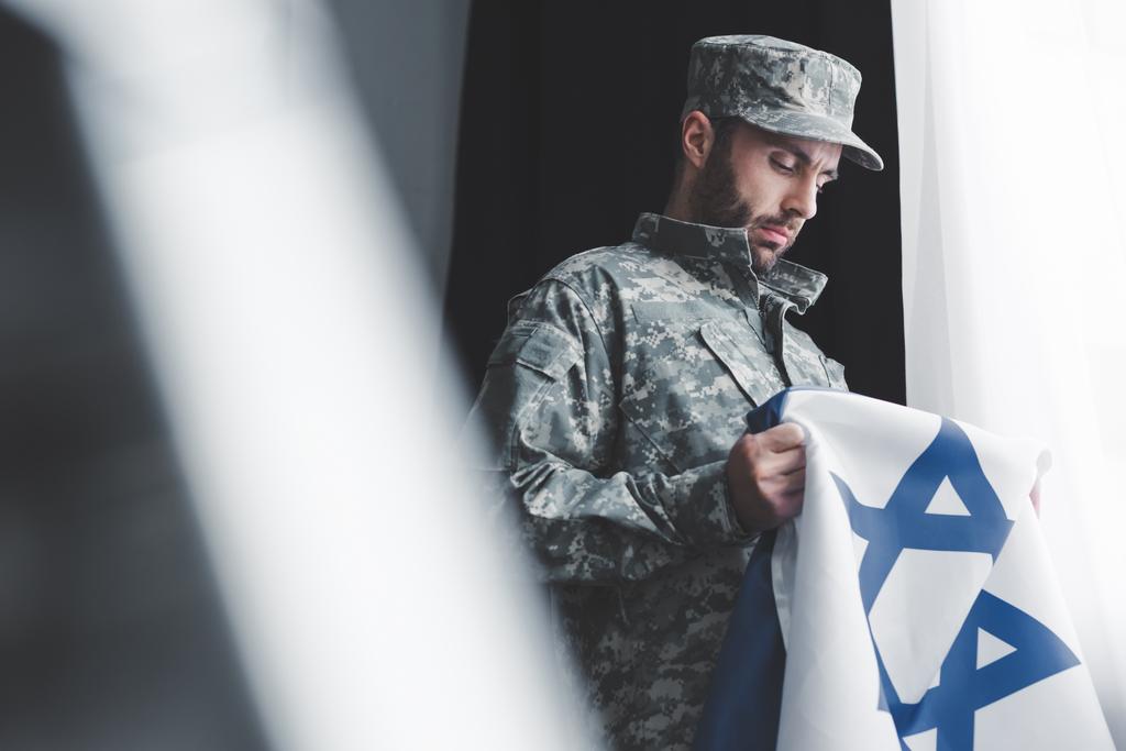 избирательный фокус вдумчивого военного в форме, держащего государственный флаг Израиля, стоя у окна
 - Фото, изображение