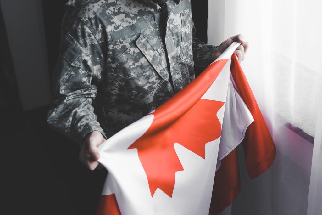 Teilansicht eines Mannes in Militäruniform, der am Fenster die kanadische Nationalflagge hält - Foto, Bild