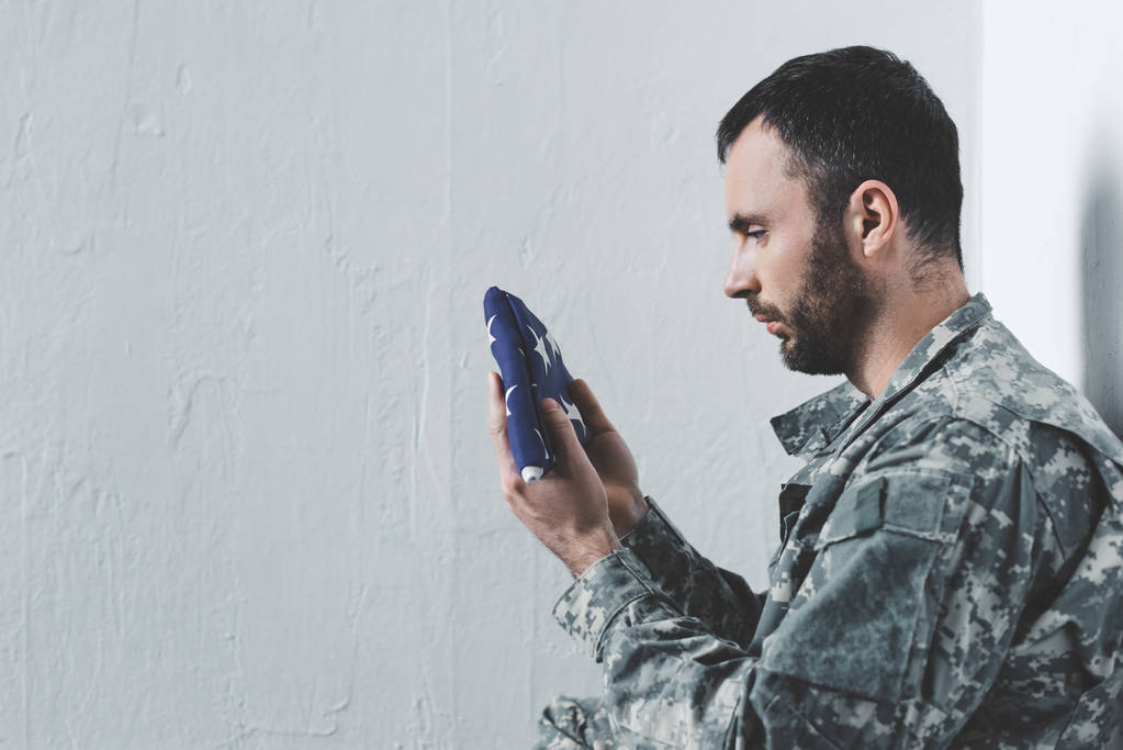 vue latérale de l'homme barbu en uniforme militaire assis près du mur blanc et tenant le drapeau national des Etats-Unis
 - Photo, image