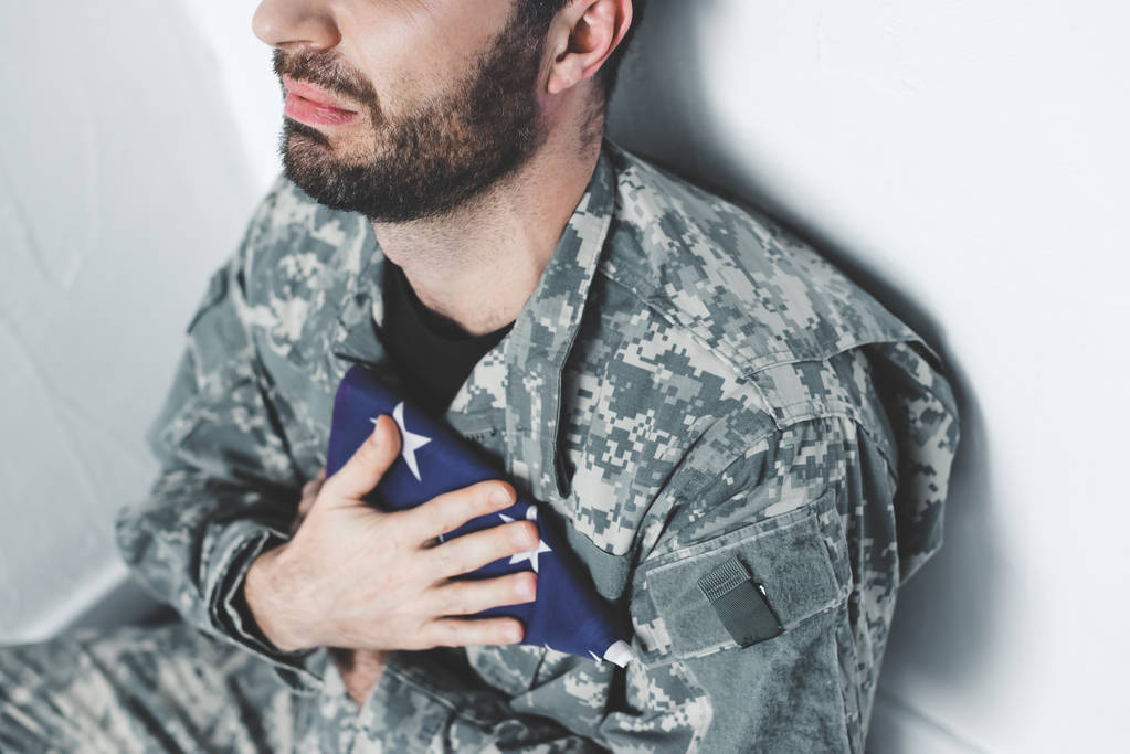 μερική θέα του καταθλιπτικός στρατιωτικός που κάθεται από το λευκό τοίχο και κρατά τις ΗΠΑ στρατιωτικό άνθρωπο κοντά στην καρδιά - Φωτογραφία, εικόνα