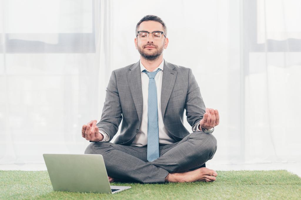 hombre de negocios sentado en la alfombra de hierba cerca de la computadora portátil y meditando en Lotus Pose
  - Foto, Imagen