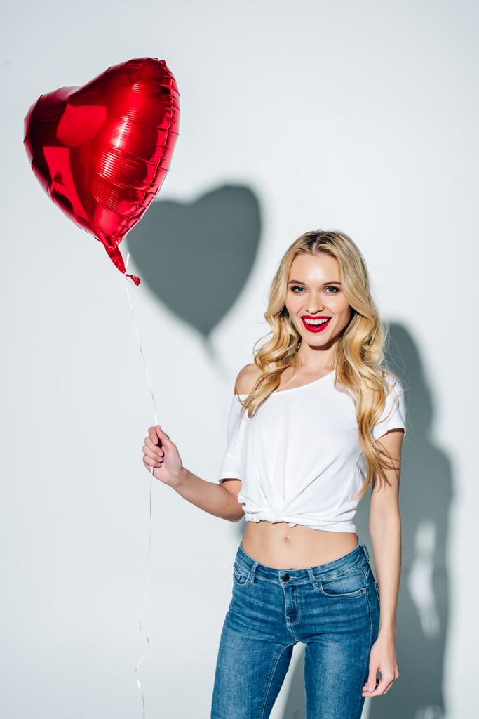 fröhliches blondes Mädchen mit rotem Ballon in Herzform, während es auf weiß steht  - Foto, Bild
