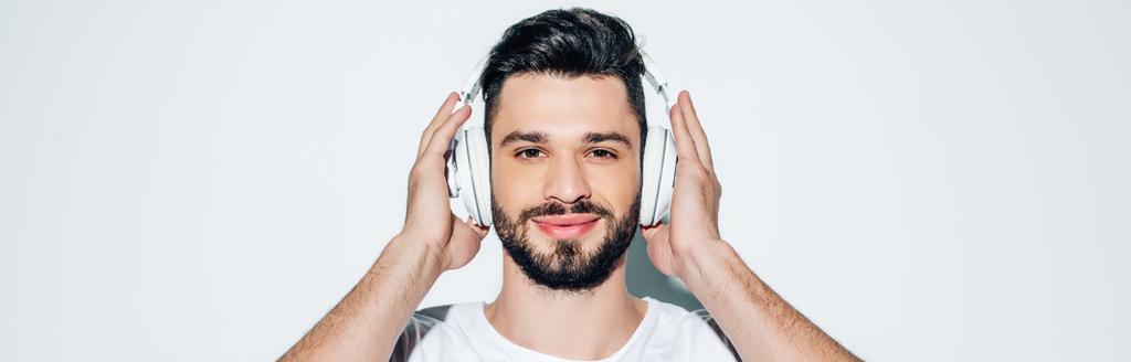 panoramiczny strzał wesoły człowiek dotykając słuchawek podczas słuchania muzyki i uśmiechając się na białym  - Zdjęcie, obraz