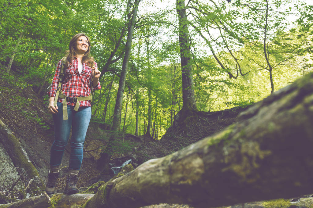 низький кут зору усміхненої молодої жінки з рюкзаком, що йде в лісі
  - Фото, зображення