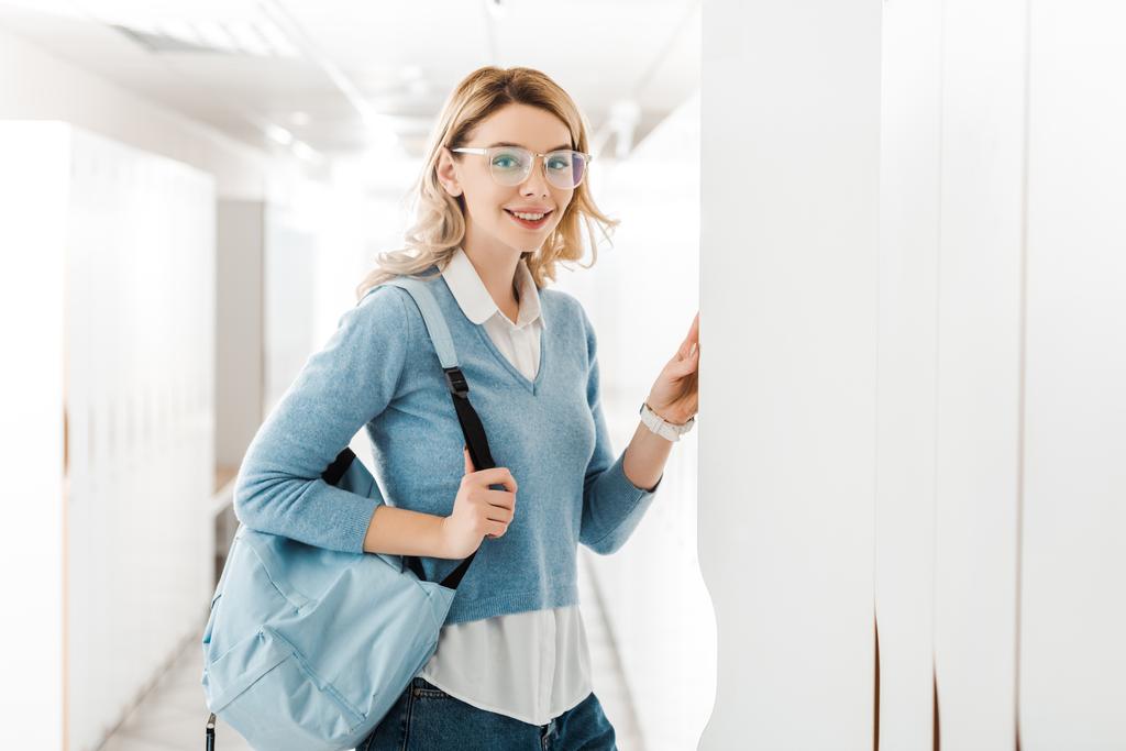 Lächelnder Student mit Brille und Rucksack öffnet Schließfach im College - Foto, Bild