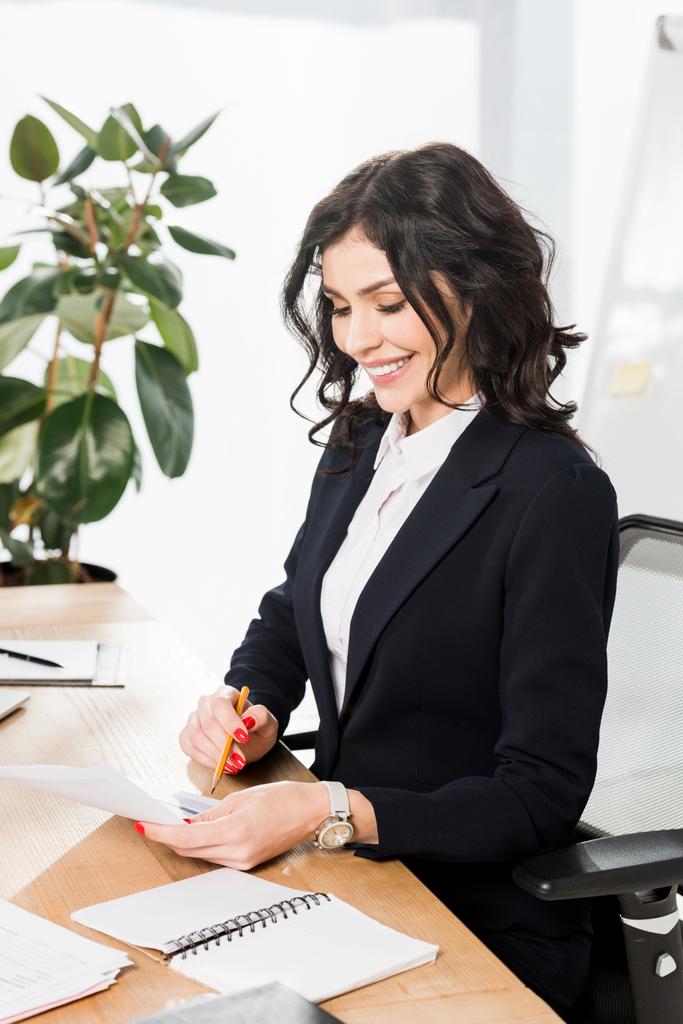 glückliche brünette Personalvermittlerin in formeller Kleidung schaut auf Dokument und hält Bleistift im Büro  - Foto, Bild