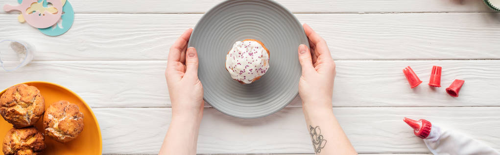 Panoramaaufnahme einer Frau, die Teller mit Cupcake auf weißem Tisch mit Zuckerguss hält - Foto, Bild
