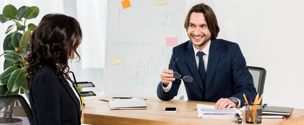 панорамный снимок счастливого рекрутера, держащего очки и смотрящего на брюнетку в офисе
  - Фото, изображение