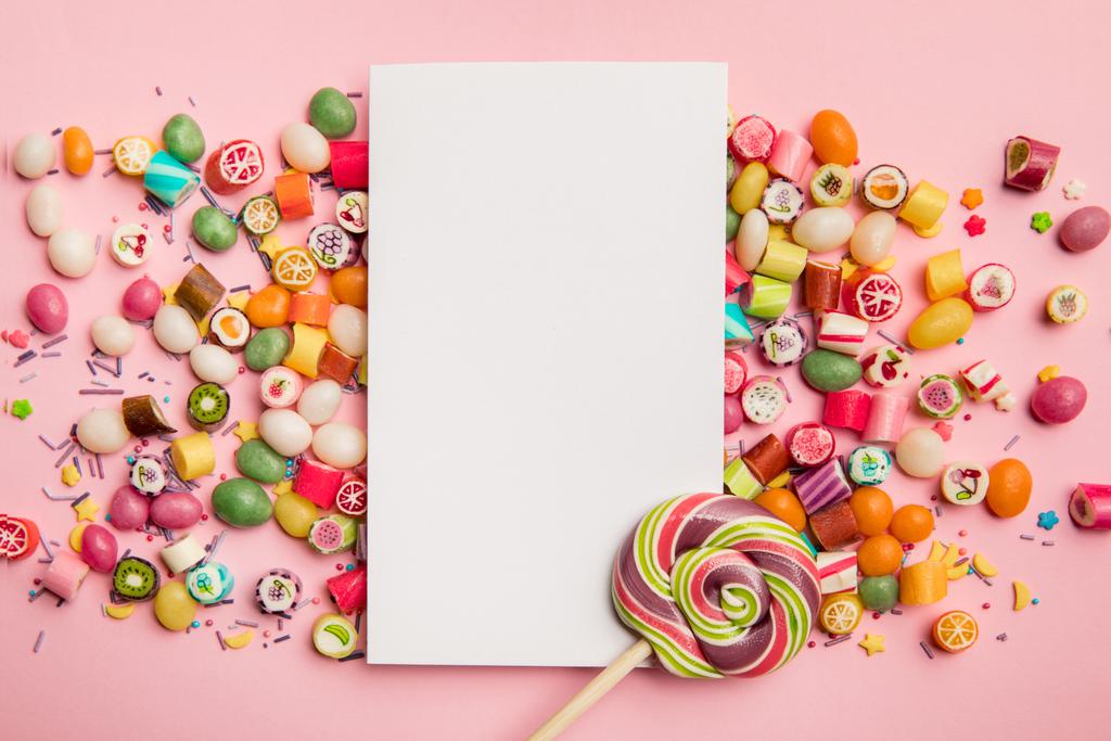 vista superior de deliciosos caramelos multicolores, piruleta y tarjeta blanca con espacio para copiar sobre fondo rosa
 - Foto, imagen