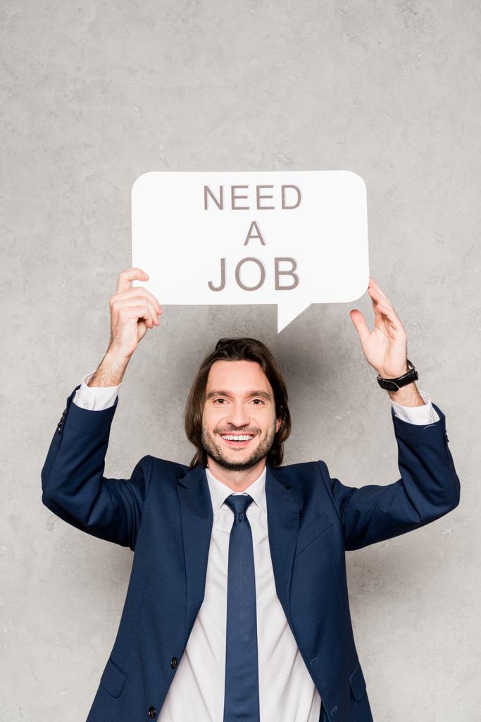 cheerful employee holding speech bubble with need a job lettering on grey - Photo, Image