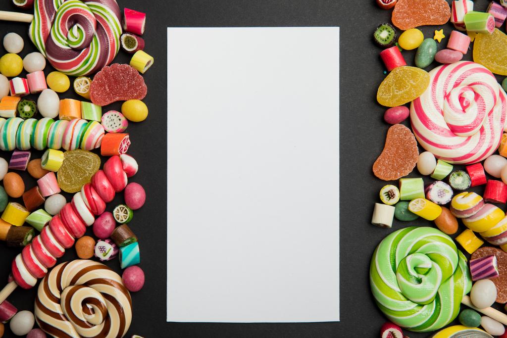 vista superior de sabrosos dulces de caramelo multicolor y piruletas alrededor de la tarjeta vacía blanca sobre fondo negro con espacio de copia
 - Foto, imagen