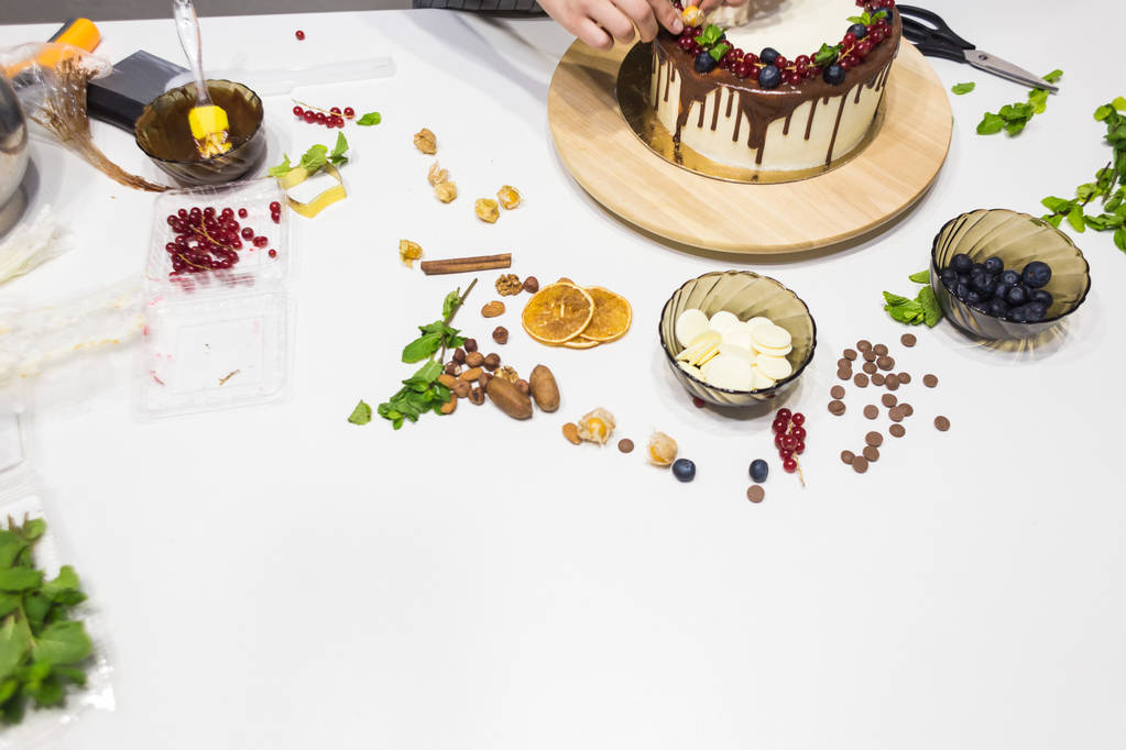 Konditor verziert mit Beeren einen Keks-Kuchen mit weißer Sahne und Schokolade. Kuchen steht auf einem Holzständer auf einem weißen Tisch. das Konzept von hausgemachtem Gebäck, Kuchen kochen. - Foto, Bild