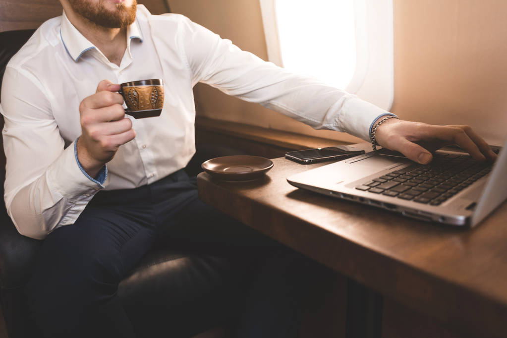 Atraktivní a úspěšný obchodník pije kávu a pracuje za laptopem, zatímco sedí v křesle svého soukromého proudového letadla. - Fotografie, Obrázek