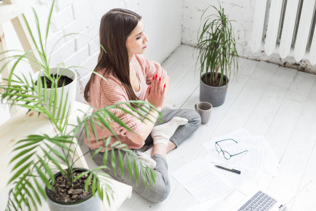 Eine junge glückliche Frau sitzt in einer hellen Wohnung in Yoga-Pose auf dem Boden und arbeitet hinter einem Laptop, ein freischaffendes Mädchen meditiert während der Arbeit. Stressabbau - Foto, Bild