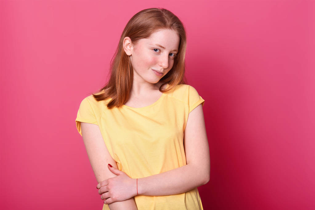 Okouzlující magnetická dívka je izolovaná přes jasně růžové pozadí ve studiu, drží ji za paži a vypadá záhadně na kameře. Talentovaný sladký model na sobě nosí světle žluté tričko. - Fotografie, Obrázek