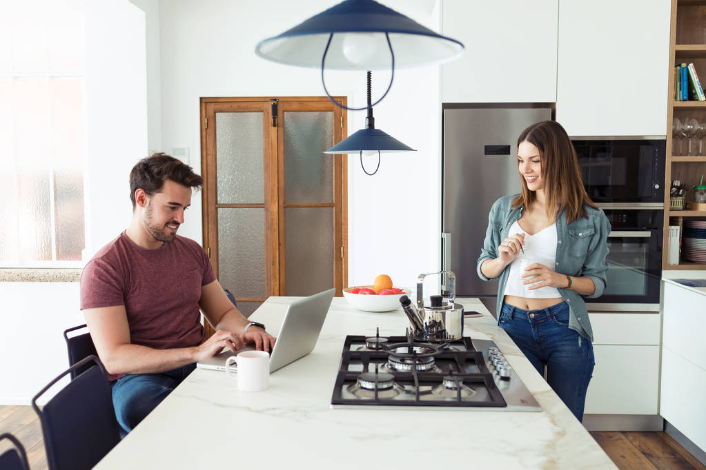 Komea nuori mies käyttää hänen kannettava tietokone ja hänen tyttöystävänsä syö jogurtti kun seisoo keittiössä kotona
. - Valokuva, kuva