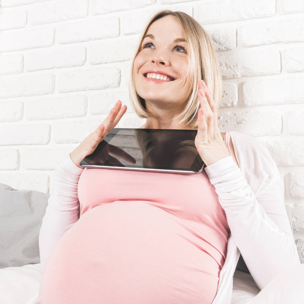 幸せな笑顔の妊娠中の女性にベッドに横し、タブレットを見て. - 写真・画像