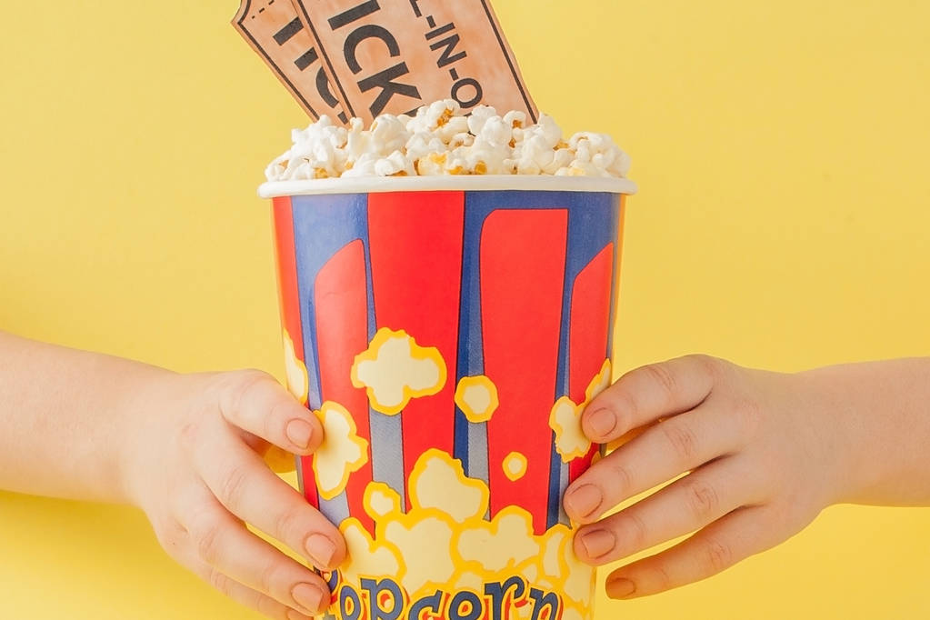 La mano prende biglietti di film e popcorn da una tazza di carta su uno sfondo giallo. Quella donna mangia popcorn. Concetto cinematografico. Piatto. Copia spazio - Foto, immagini