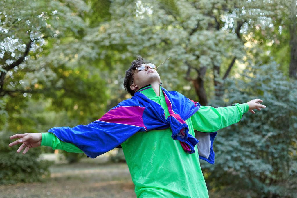 Young handsome caucasian man with bright freaky sportswear in 70s style stands at a morning park, hands widely outstretched. Stick in the mouth, golden glasses, extravagant fashion. Outdoors, copy space. - 写真・画像