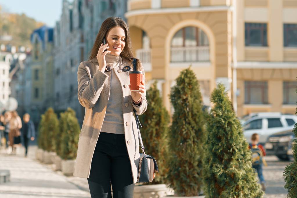 junge lächelnde schöne Frau, die auf der Straße einer sonnigen Herbststadt läuft und mit dem Handy telefoniert - Foto, Bild