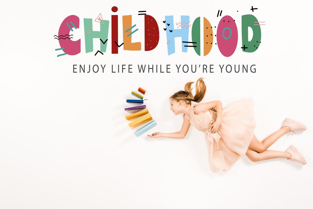 Widok z góry wesoły dzieciak w różowej sukni trzyma książki i pływające w pobliżu dzieciństwa cieszyć się życiem, podczas gdy jesteś młody napis na białym - Zdjęcie, obraz