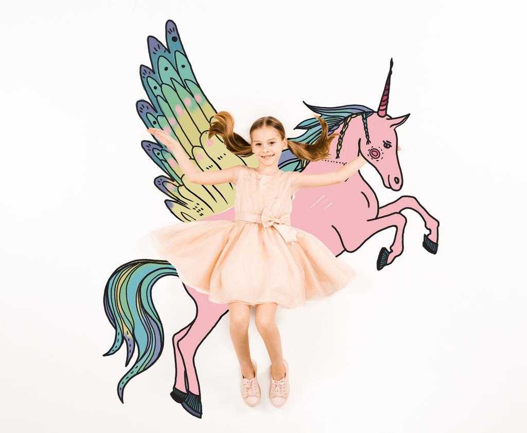 top view of cheerful kid looking at camera and gesturing near unicorn on white  - Photo, Image