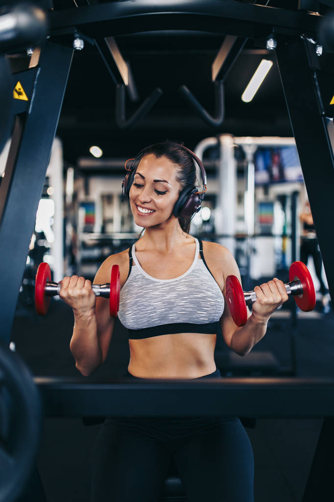 Jeune femme en forme et séduisante travaillant dans une salle de gym moderne et écoutant de la musique avec casque bluetooth et téléphone intelligent
. - Photo, image