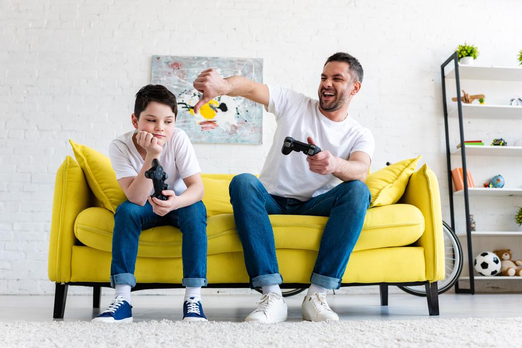 isä osoittaa peukalo alas, kun istuu pojan kanssa ja pelaa videopeliä kotona
 - Valokuva, kuva