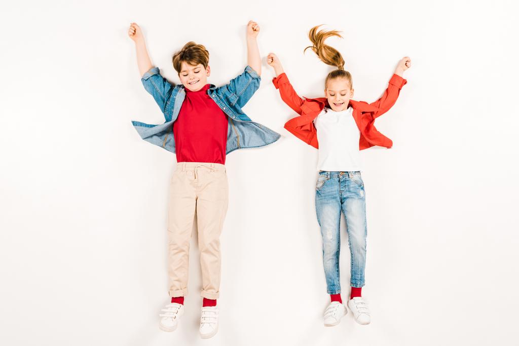 Draufsicht auf fröhliche Kinder mit Händen über dem Kopf, die auf weiß liegen  - Foto, Bild