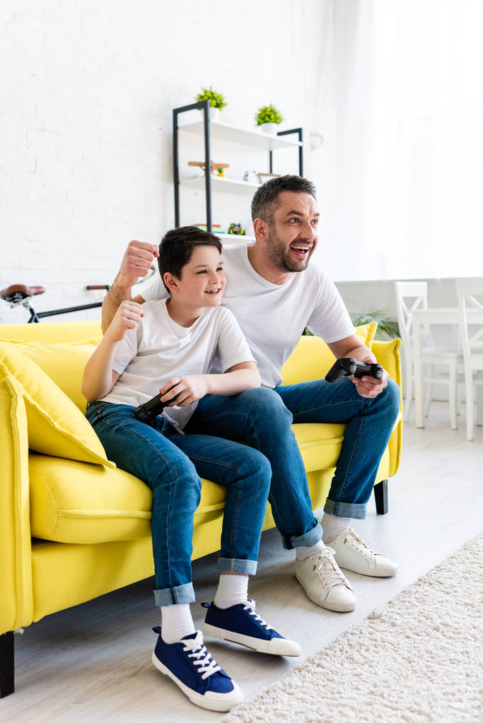 ενθουσιασμένος πατέρας και γιός επευφημίες ενώ παίζοντας βίντεο παιχνίδι στον καναπέ στο σπίτι στο σαλόνι - Φωτογραφία, εικόνα