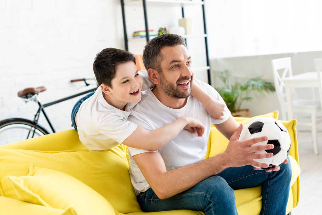 щасливий батько і син дивиться спортивний матч на дивані з футбольним м'ячем вдома
 - Фото, зображення
