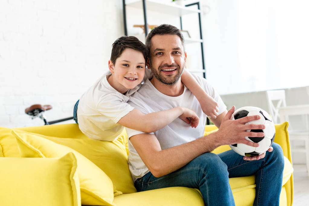 счастливые отец и сын на диване с футбольным мячом смотреть в камеру дома
 - Фото, изображение